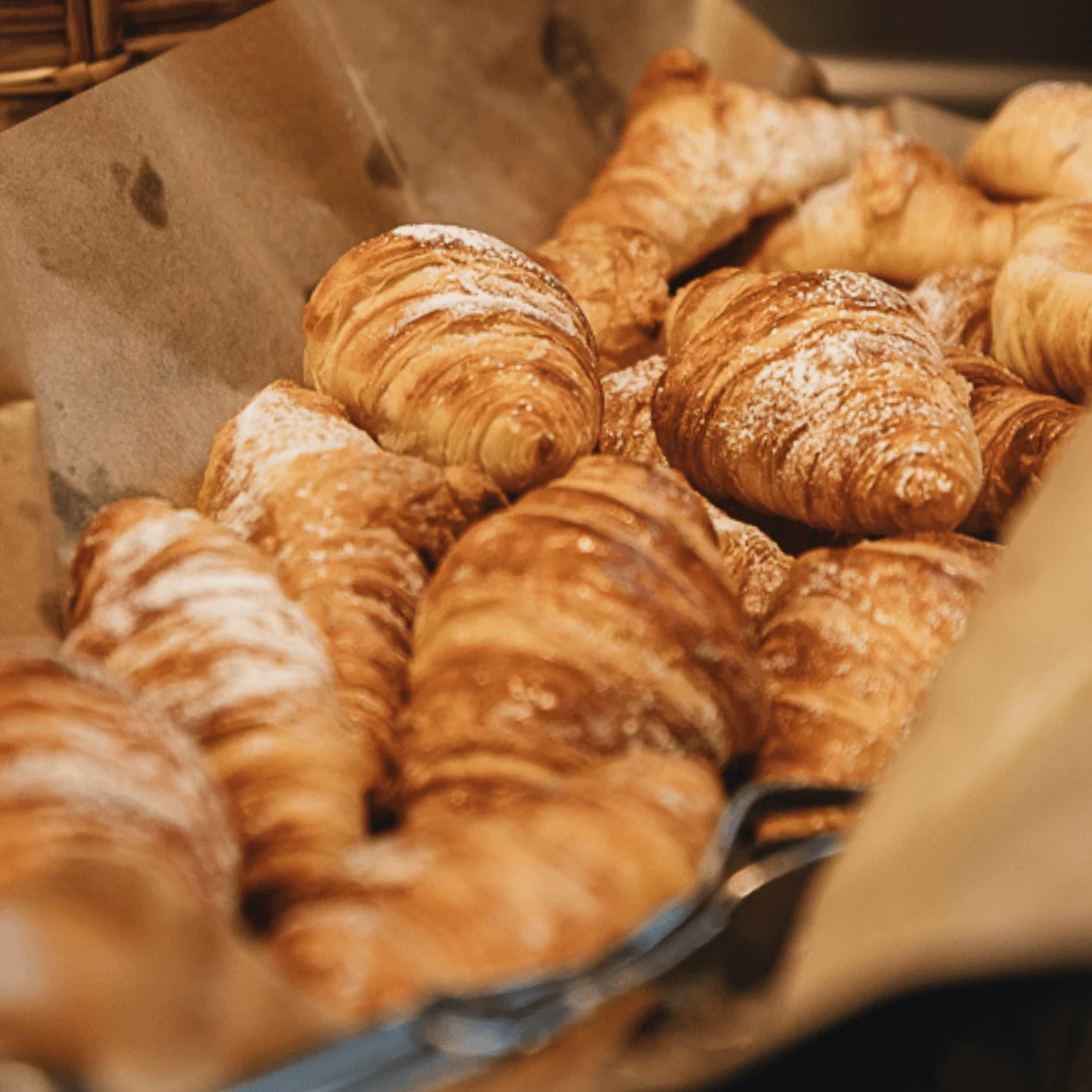 Croissanterna är staplade, beströdda med florsocker, på brunt papper. De ligger i en korg och skapar en mysig bageriatmosfär.