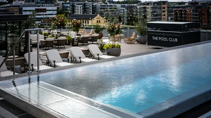 A rooftop pool with clear, calm water; surrounded by white lounge chairs and plants in pots. Nearby, a sign reads "THE POOL CLUB," and apartment buildings are in the background.