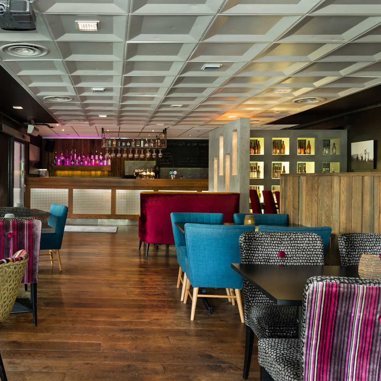 Bar med färgglada stolar omger bord; bardisk i bakgrunden med flaskor och glas; varm belysning förstärker den mysiga, moderna atmosfären.