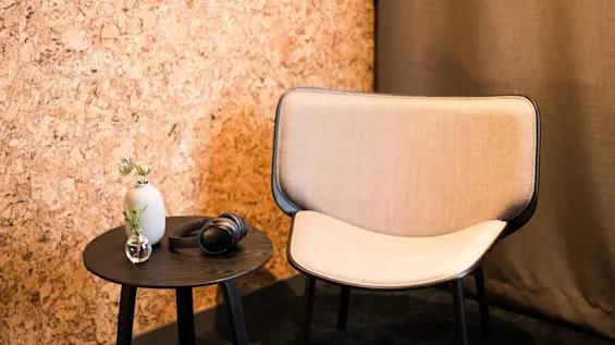 A beige chair sits next to a small round black table with headphones and plants. Cork wall with three small artworks hangs nearby, alongside a brown curtain.