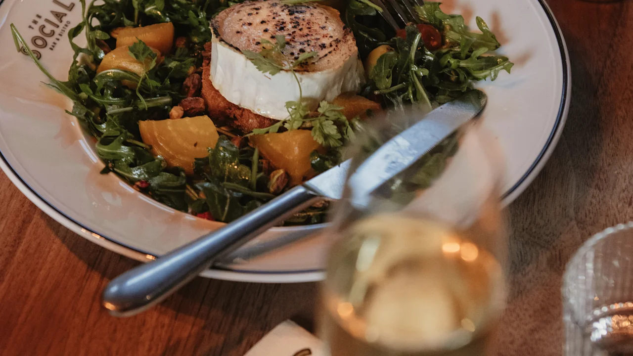 A plate of fresh arugula salad with goat cheese, yellow beets, and walnuts sits on a wooden table, accompanied by a glass of white wine. Text: "THE SOCIAL WINE BISTRO."