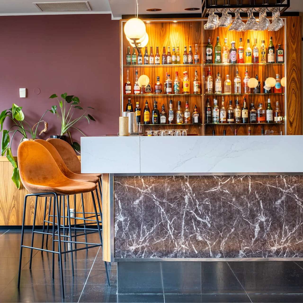 A marble-topped bar with two brown stools stands before a backlit shelf displaying various bottles. A plant sits nearby against a purple wall, creating a modern setting.