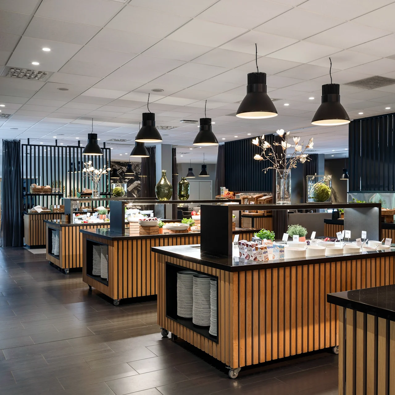 Buffet tables display diverse foods in an elegant, modern dining area. Overhead lights illuminate the neatly arranged dishes, creating an inviting atmosphere with sleek black and wooden accents.