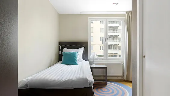 A bedroom with a striped bed, teal pillow, and nightstand by a window overlooking other buildings.
