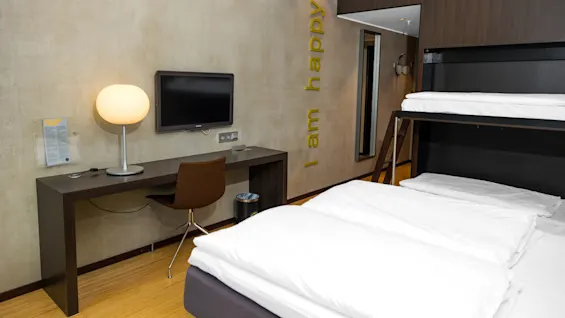 A hotel room includes a desk with a lamp and TV, bunk bed, and wall decoration reading "I am happy," against a beige wall.