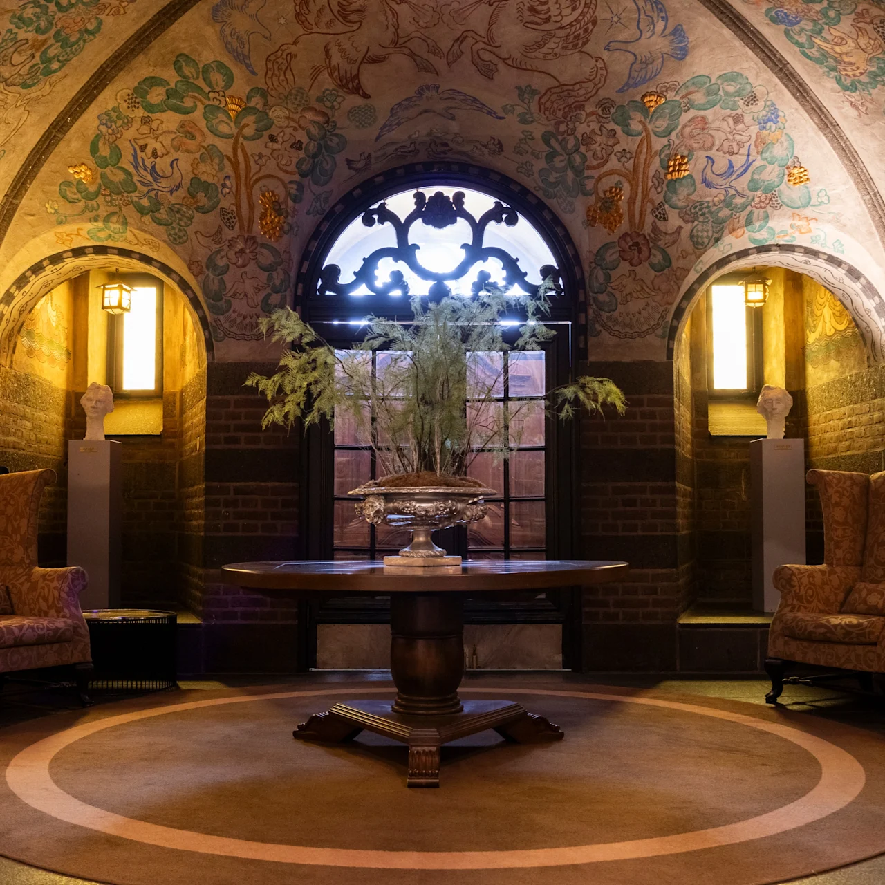Elegant round table holding a decorative centerpiece; flanked by vintage armchairs in a vaulted room with intricate ceiling art and warm lighting from wall sconces, creating a cozy, luxurious atmosphere.