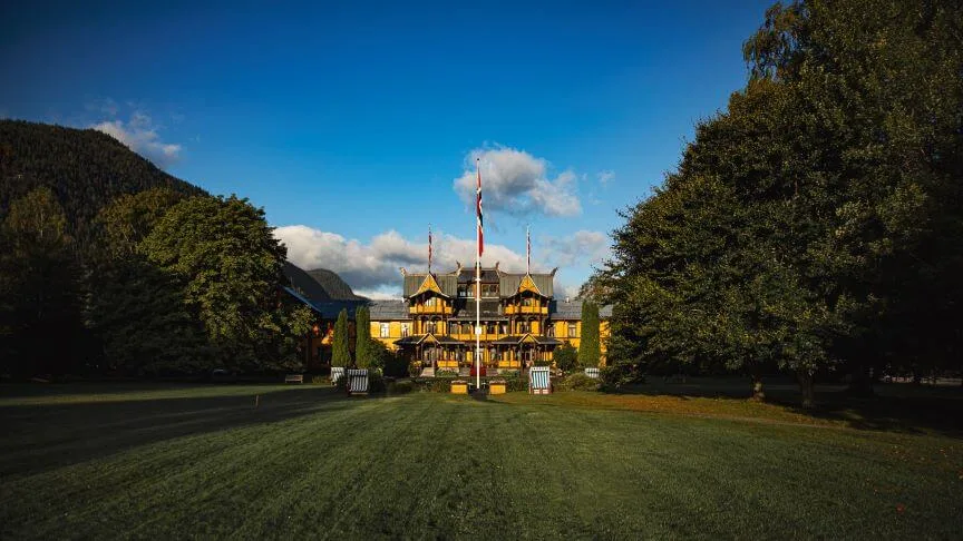 A grand wooden building stands amid lush greenery, bathed in sunlight, with a flagpole in the foreground. Mountains and a blue sky provide a scenic backdrop.