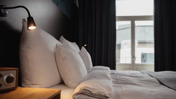 A neatly made bed with two pillows is illuminated by bedside lamps in a dimly lit room. Thick curtains partly cover a window looking out onto a building.