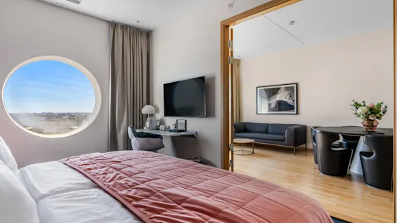 En moderne hotelsuite med et soveværelse med et rundt vindue og et separat opholdsområde med sofa og spisebord.