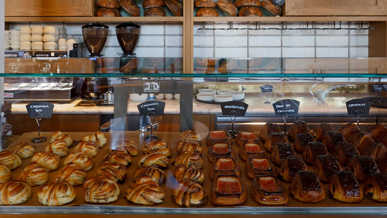 Bagværk pænt arrangeret bag en glasskærm, nogle markeret "Chaussons aux Pommes", "Pain au Chocolat". Hylder med brød i baggrunden giver en varm, indbydende bageriomgivelse.