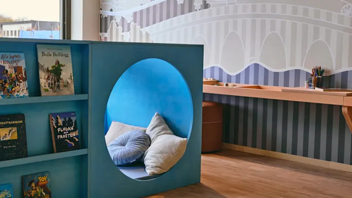 Et fargerikt lekeområde for barn med en blå bokhylle, en koselig lesekrok og et håndverksbord. Bøker inkluderer Bulle Bulldogg, Flugan Frosten og Toy.