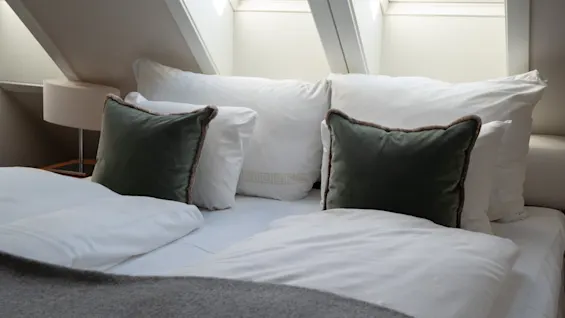 En koselig soveromsscene med en pent redd seng, hvitt sengetøy, mørkegrønne puter og et grått pledd. Takvinduer over.