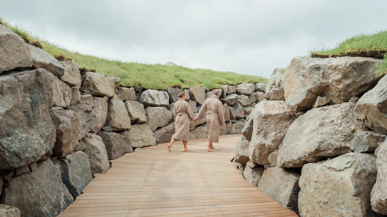 Two people in bathrobes walk hand-in-hand on a wooden path between rock walls and grassy hills.