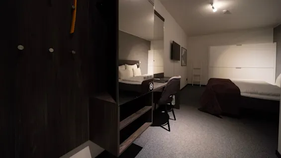 Et hotelværelse har en seng med puder, et mørkt tæppe, et TV på væggen og et skrivebord med en stol. Tekst på spejlet siger, "YOU LOOK GREAT." Et "rygning forbudt"-symbol er også synligt.