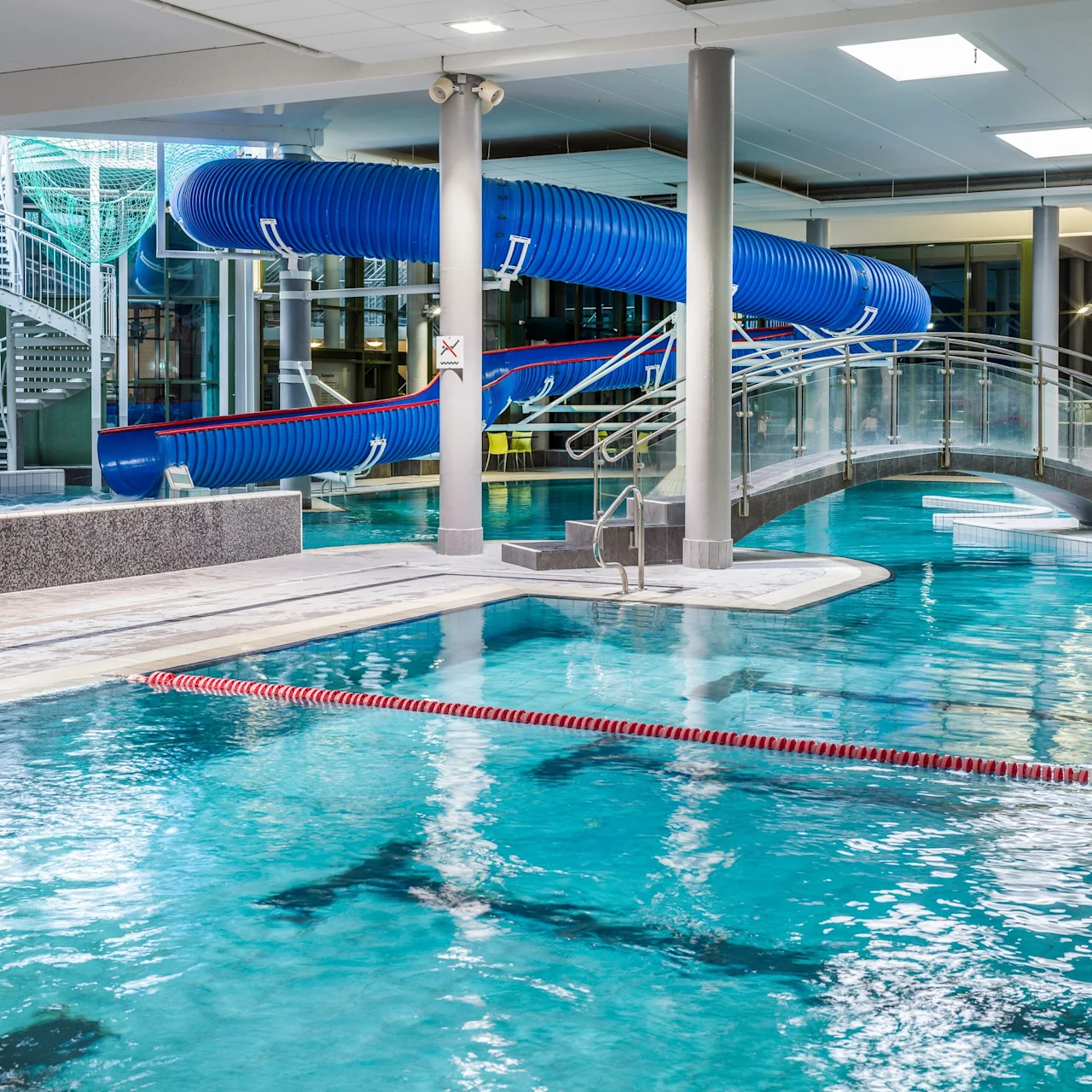 A large indoor swimming pool features a blue water slide and a small footbridge. The surrounding area includes columns, a spiral staircase, and seating, creating a recreational environment.