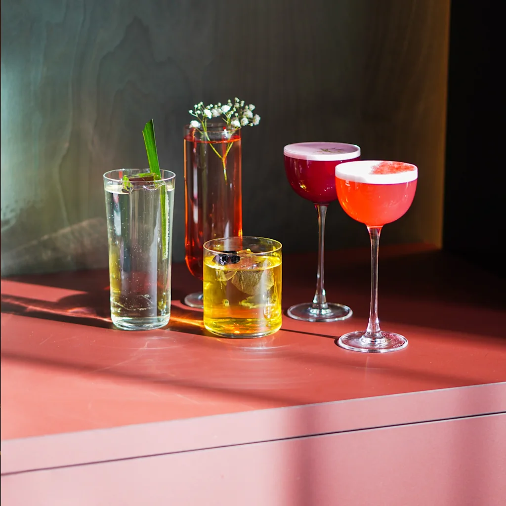 En samling fargerike cocktails og drinker på en rødbrun overflate, med mørk bakgrunn og flekkete sollys.