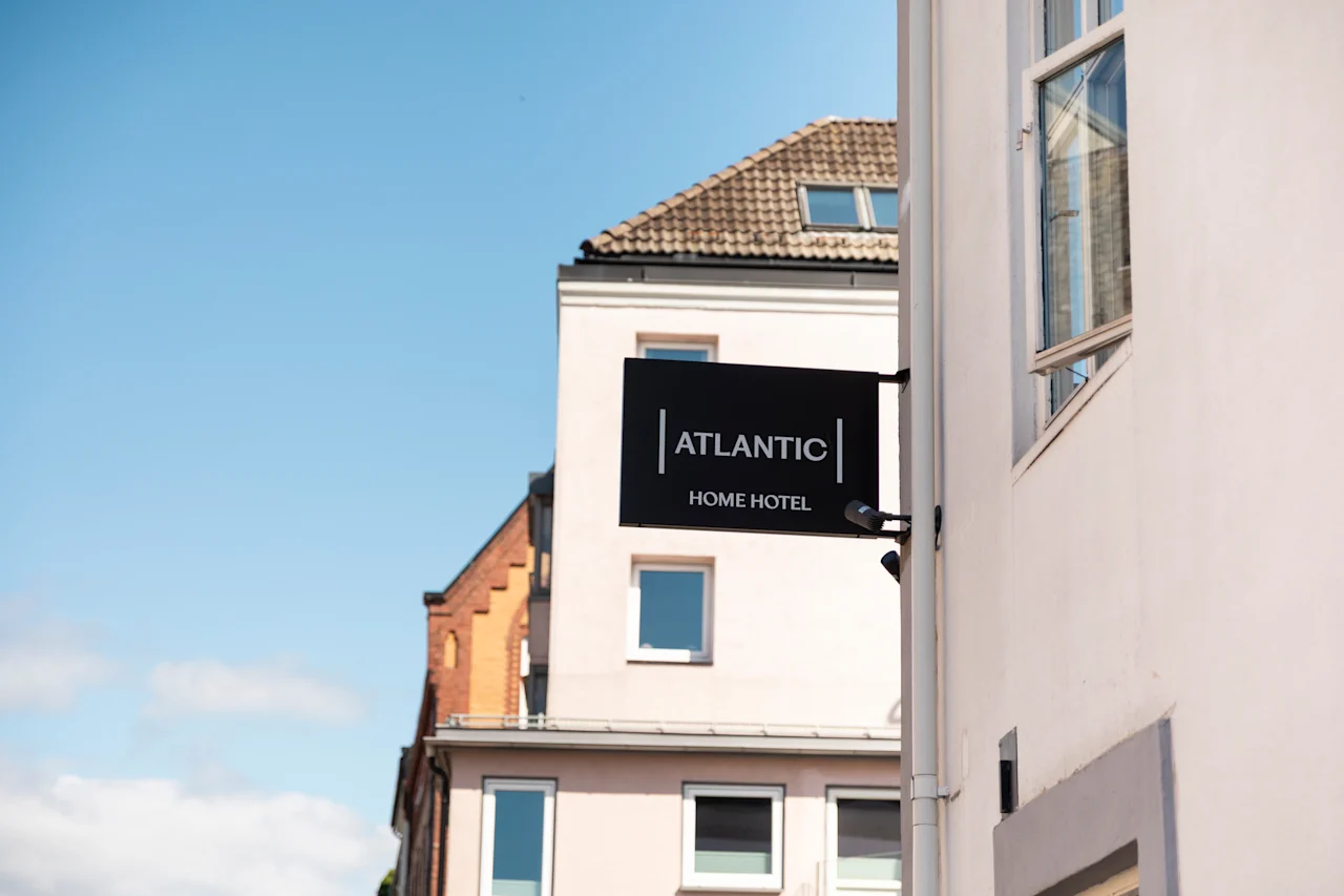 Et sort skilt med teksten HOME HOTEL er fastgjort til en hvid bygning, med et glimt af andre strukturer og en blå himmel.