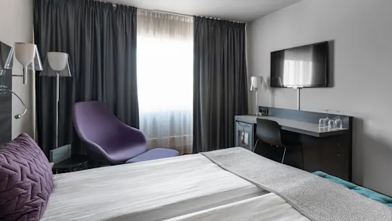 En säng med lila kuddar är prydligt bäddad bredvid ett fönster med mörka gardiner. I närheten finns en lila stol och ett skrivbord med glas och en TV i ett hotellrum.