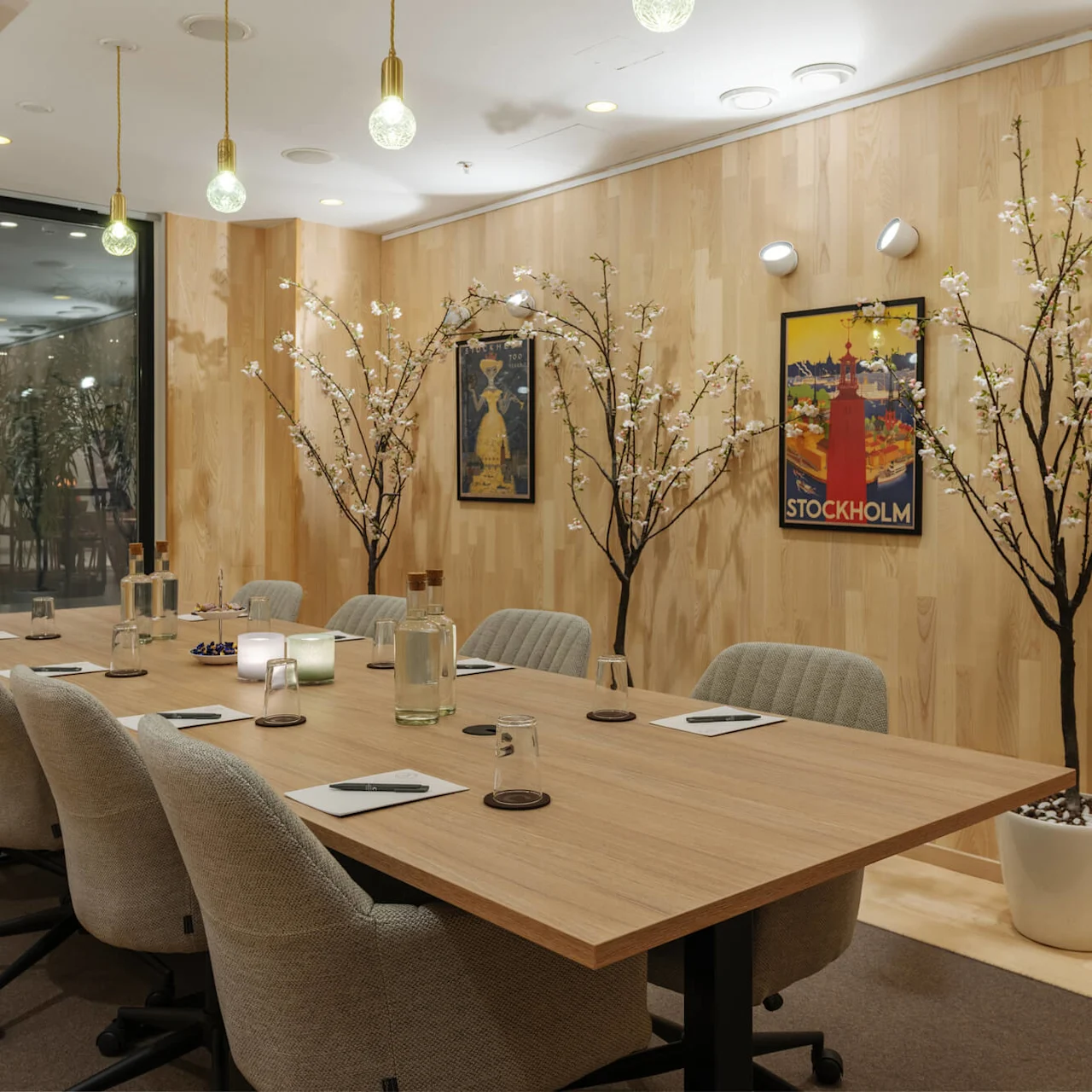 A meeting room features a wooden table surrounded by grey chairs, set with notepads and water bottles. Faux cherry blossom trees adorn the walls, alongside colorful Stockholm posters.