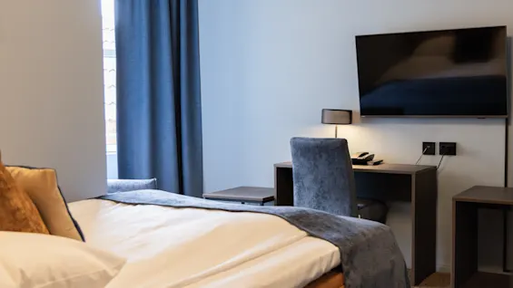 A neatly made bed with pillows sits near a window with blue curtains. A desk with a chair and a wall-mounted TV complete the modern room setting.