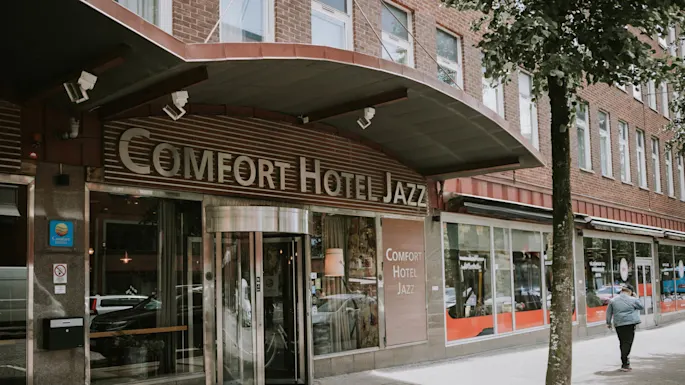 Et hotellinngang med en roterende dør og et skilt som leser "COMFORT HOTEL JAZZ" i et urbant miljø.