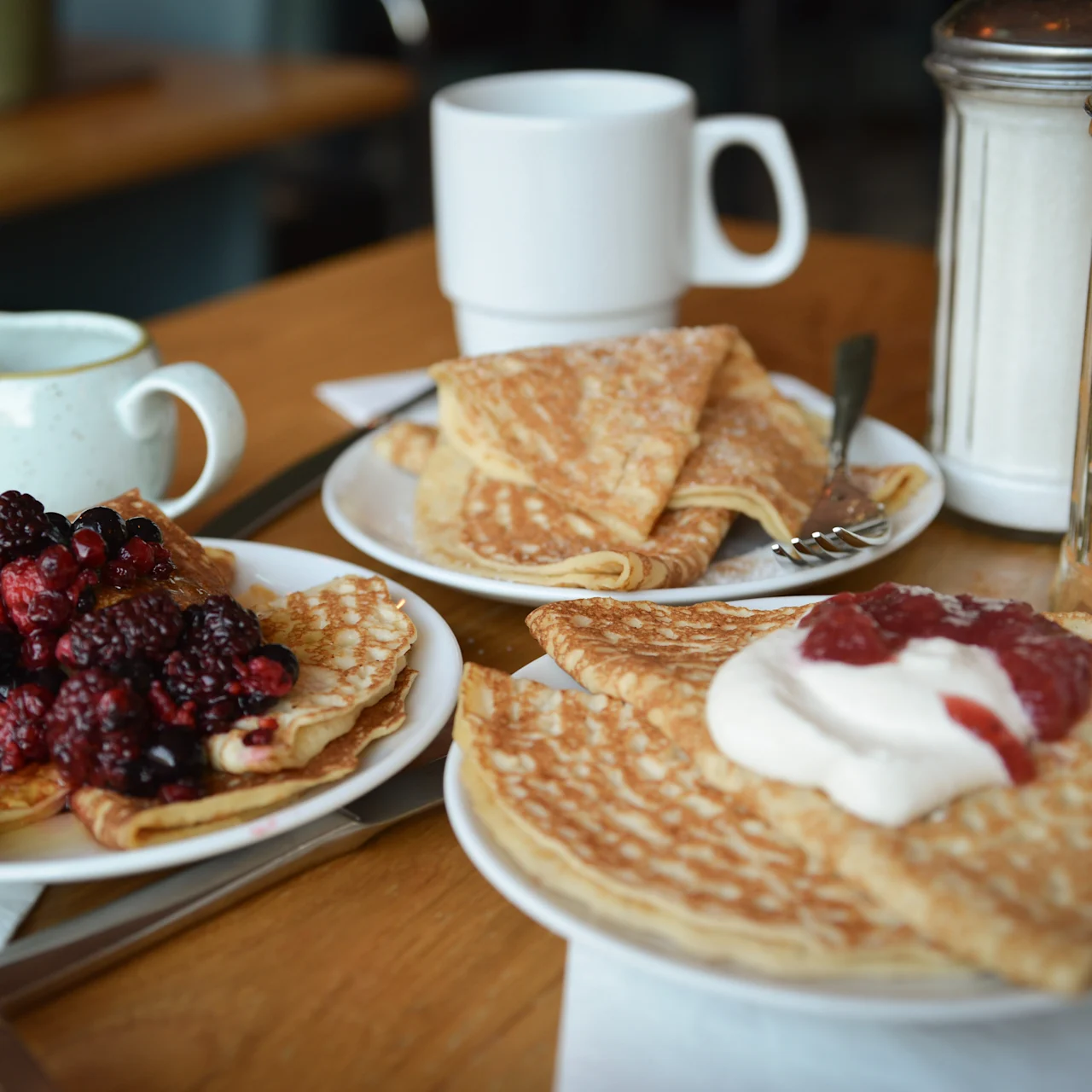 Pandekager toppet med bær og flødeskum på tallerkener med gafler på et træbord. Omgivende genstande inkluderer kaffekopper, en flødekande og sukkerstrøere, hvilket antyder en morgenmadsscene.