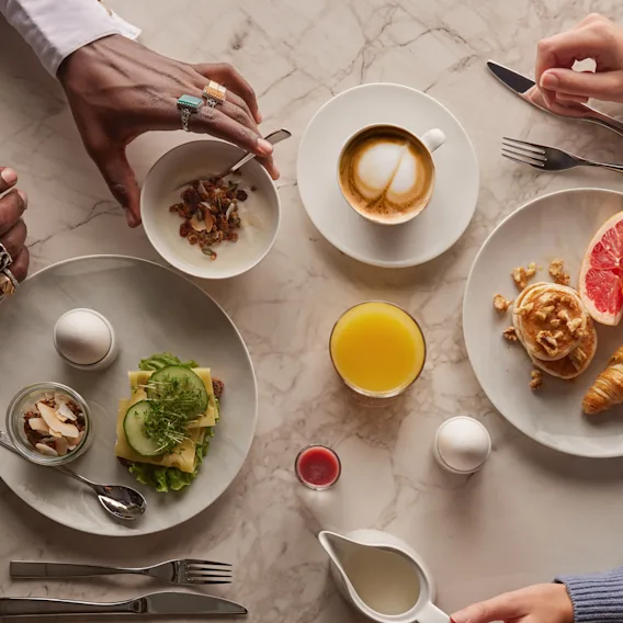En marmorbord med händer som arrangerar frukostartiklar. Tallrikar innehåller en smörgås, ägg, sallad, yoghurt, croissant, grapefrukt och en våffla. Drycker inkluderar kaffe, juice och en röd dryck.