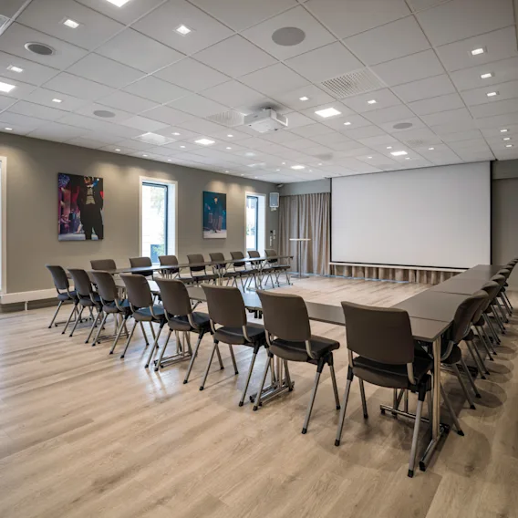 Et moderne konferanserom med U-formet bordoppsett, stoler, stor prosjektorskjerm og vinduer, klart for et møte.