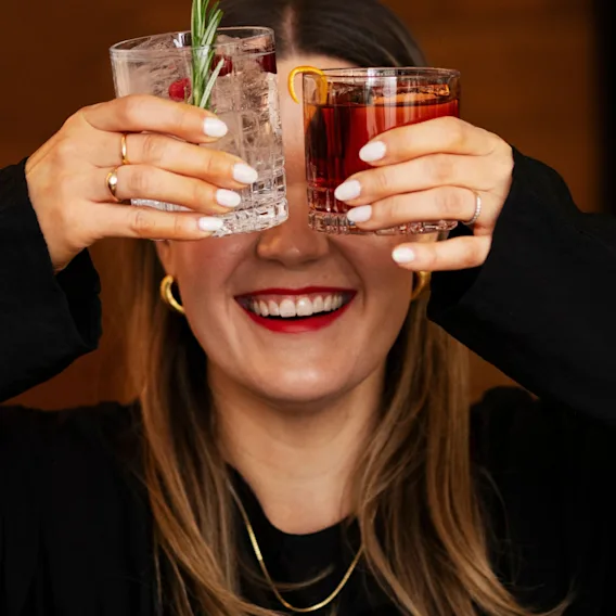 En person med långt hår håller två cocktails framför ansiktet, gömmer det. Ett glas har en klar vätska och rosmarin; det andra har en röd dryck. Bakgrunden är suddig.