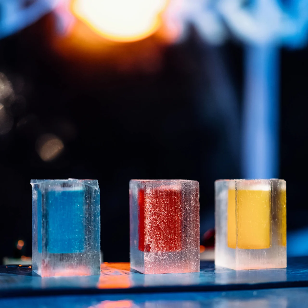Three translucent ice cubes with blue, red, and yellow centers stand on a reflective surface, illuminated by a blurred, warm light in a dark background.