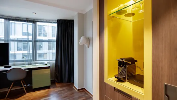 A coffee machine sits on a yellow shelf, surrounded by glasses. Nearby, a workspace with a chair and desk faces large windows covered by sheer curtains.