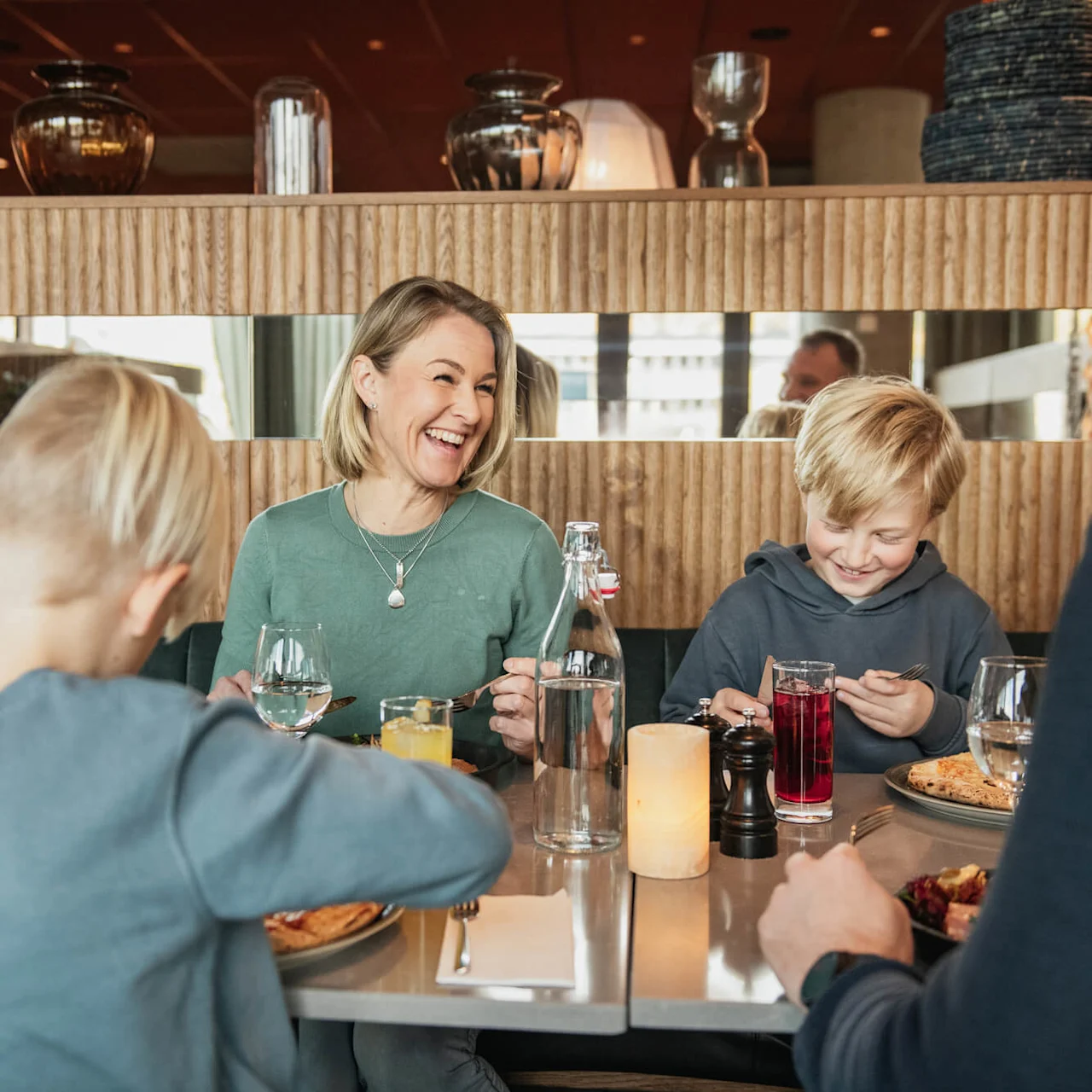 En familj sitter runt ett restaurangbord, ler och njuter av mat. Drycker och tallrikar finns på bordet; bakgrunden har träinredning och speglar.