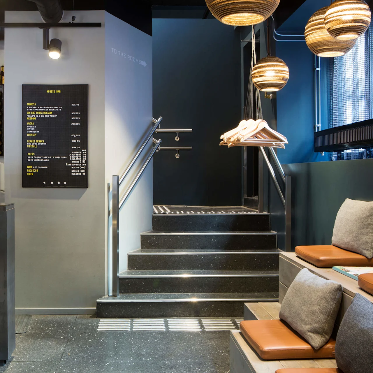 A bar counter displays bottles above shelves. To the right, stairs lead up, flanked by hanging lights and chairs. A menu lists drinks: MIMOSA, GIMLET, NEGRONI, APEROL SPRITZ, WHISKEY, FINNISH, BEERS, WINE, PROSECCO, CIDER.