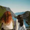 To personer som sitter og ser utover en vakker, dypblå fjord omgitt av høye, skogkledde fjell.