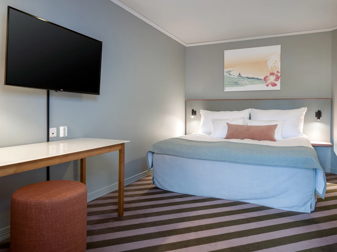 En seng med hvitt og blått sengetøy er plassert i et hotellrom. Et maleri henger over sengen. Et bord og en krakk står overfor, med en flatskjerm-TV på veggen.