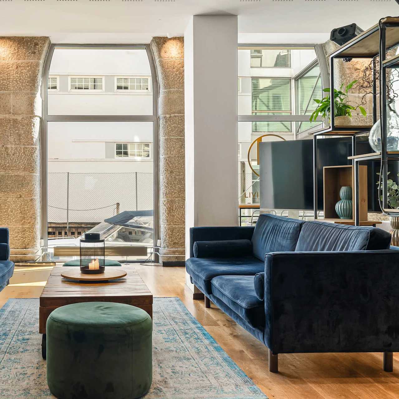 To mørkeblå sofaer står overfor hinanden i en moderne stue med store vinduer, lyst trægulv og et centralt sofabord med et lys. En plante pynter en hylde.