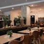 A cozy restaurant interior with wooden tables, leather chairs, and green plants. Hanging lights illuminate the space. A chalkboard reads THE TASTE OF HOME.