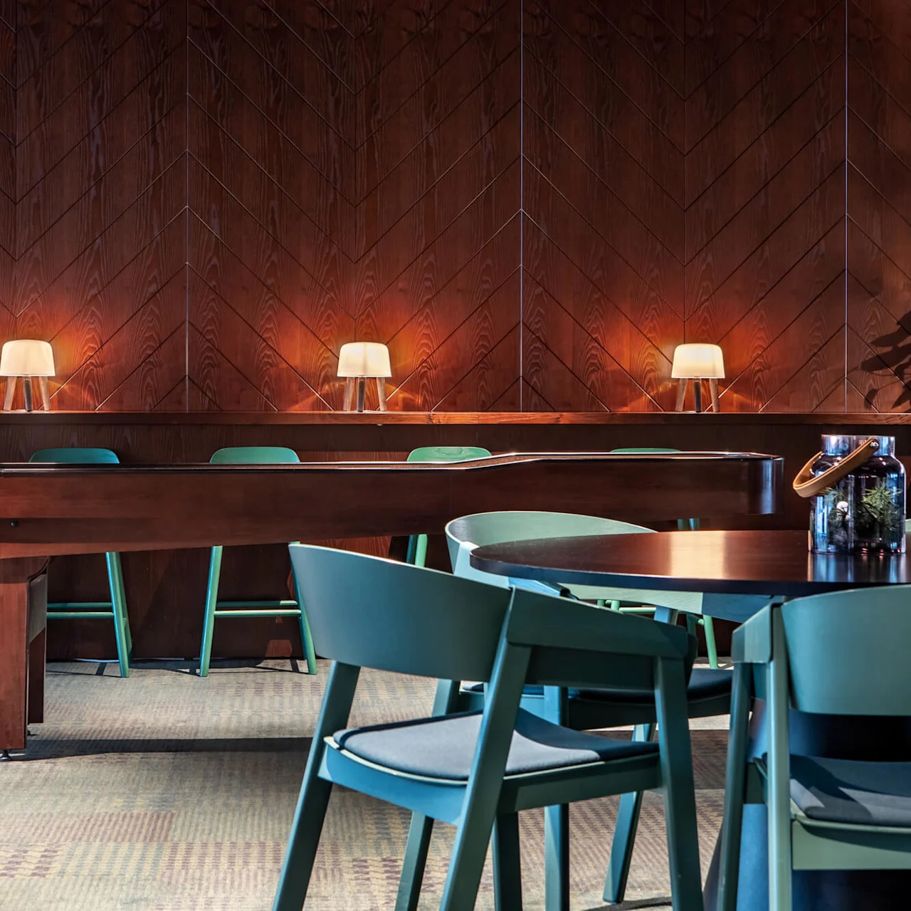 A row of modern lamps illuminates green chairs against a wooden wall, with a circular table and chairs in the foreground, creating a cozy dining area ambiance.