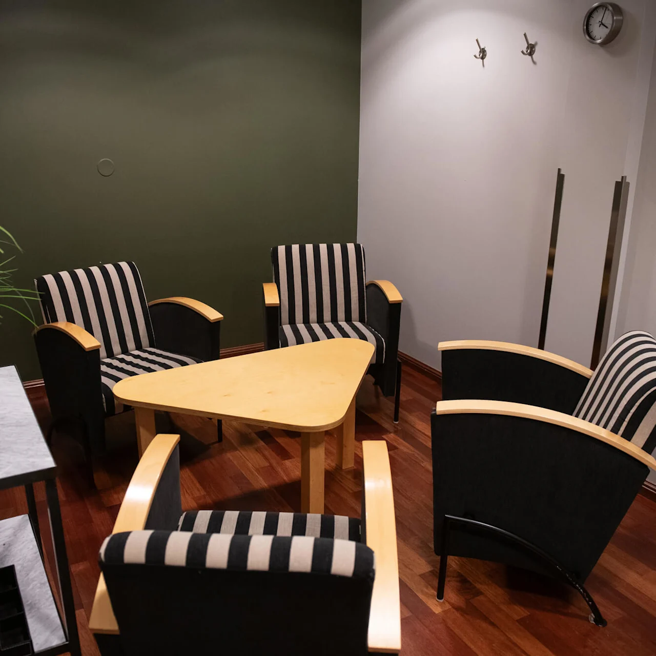 Four striped armchairs encircle a triangular wooden table in a small room. A black vase with green plant sits on a side table. Wooden floor and wall clock enhance the setting.