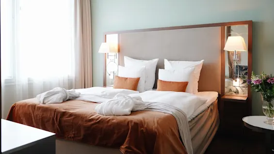 A neatly made bed with pillows and brown blanket, flanked by bedside lamps, in a softly lit, elegant bedroom featuring a vase of flowers on a side table.