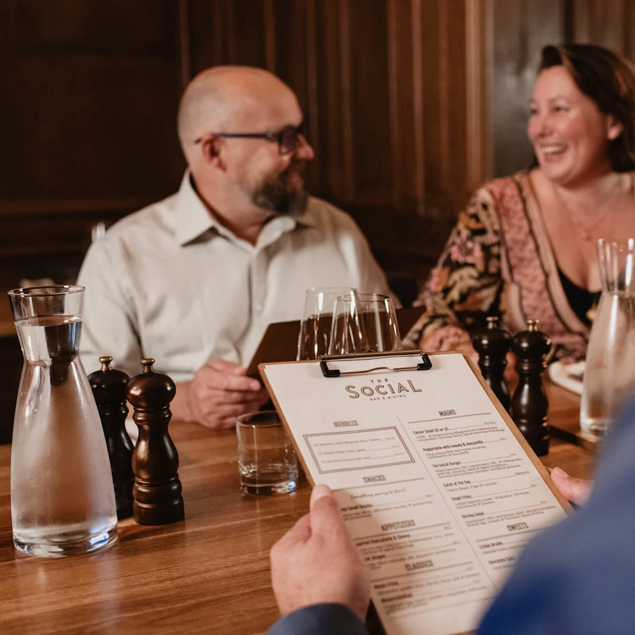 To personer spiser middag, en holder en meny med tittelen "THE SOCIAL", i en koselig restaurantinnstilling.