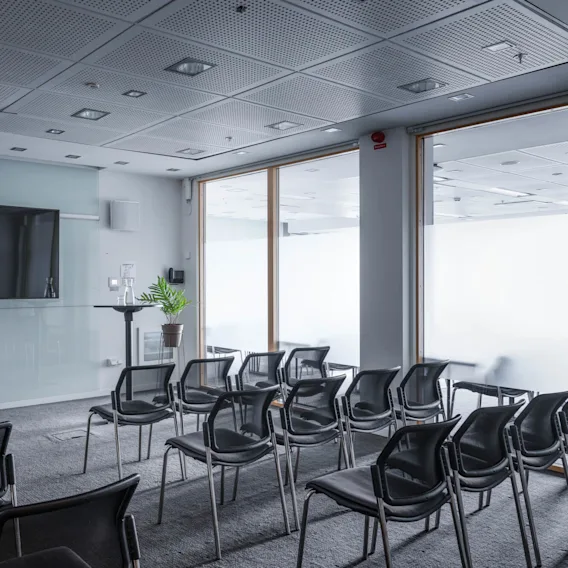 A row of black chairs faces a large screen in a conference room. The room has a grey carpet, glass walls, and a potted plant near the screen.