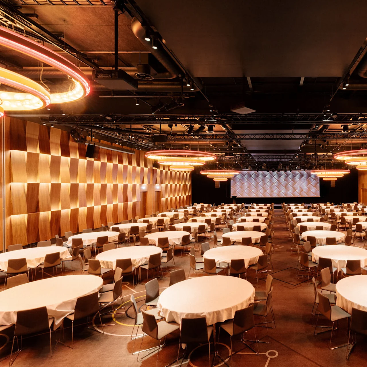 Round tables with white tablecloths are set in rows, surrounded by chairs, in a large conference hall. Soft lighting fixtures hang from the ceiling, creating a warm ambiance.