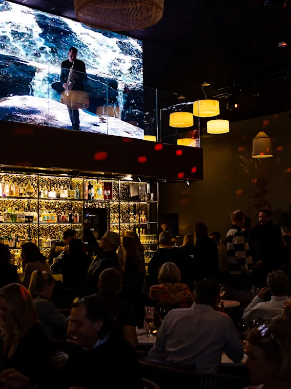 En bar är full av människor som umgås under varma ljus. Ovanför visas oceanbilder på en stor skärm. Hyllor bakom baren är fyllda med flaskor och skapar en livlig atmosfär.