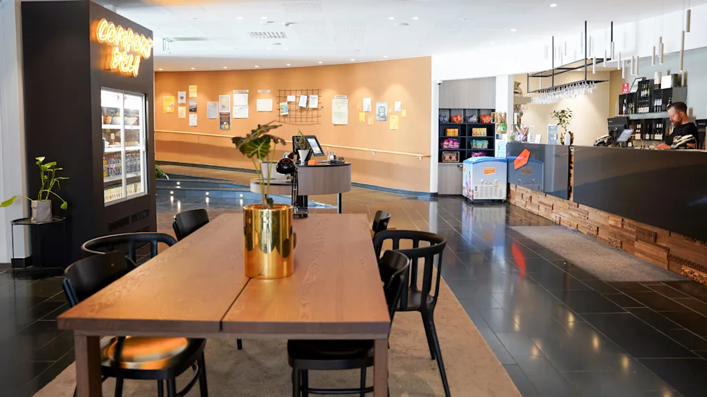 A café interior with wooden tables, black chairs, and a counter. Neon sign reads "Corner's Deli," plants accent the space, and customers are present.