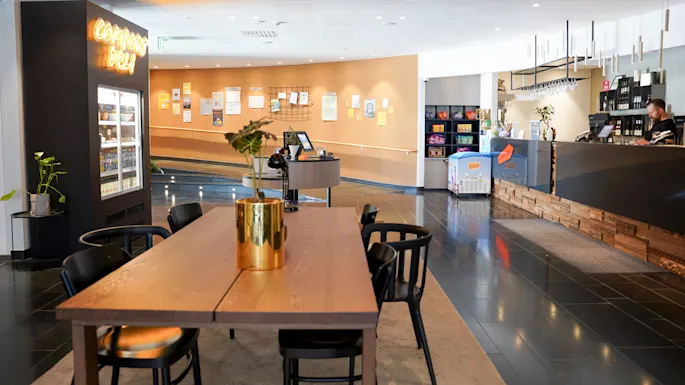 Caféinteriør med trebord, sorte stoler og en disk. Neonskilt med "Corner's Deli", planter som dekor og kunder tilstede.