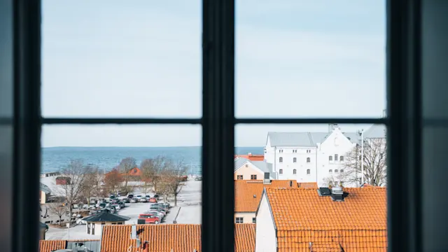 Udsigt gennem et vindue; orange-taget bygninger og parkerede biler i forgrunden. Træer og en stor vandmasse i det fjerne under en klar himmel.