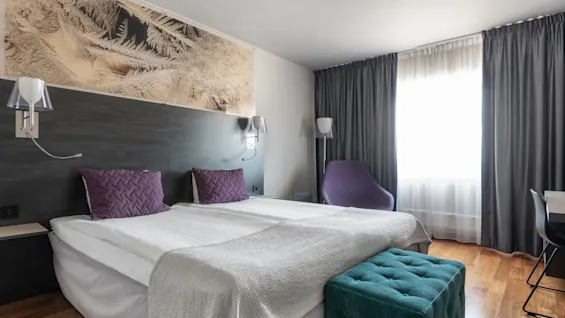 Ett modernt hotellrum har en stor säng med vita sängkläder och lila kuddar. Ett väggkonstverk och mjuk belysning framhäver den samtida inredningen, med ett fönster täckt av grå gardiner i närheten.