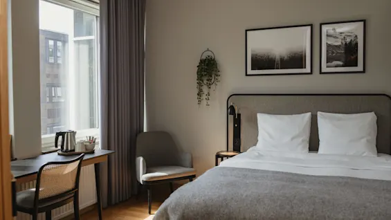 En pent senger med hvite puter og grått sengetøy flankert av innrammet kunst, en stol, og et skrivebord med vannkoker, i et mykt opplyst rom.