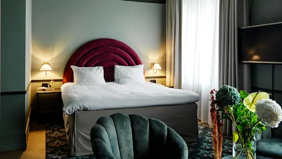 A luxurious bed with a red, semi-circular headboard is flanked by bedside lamps in a stylish, light-filled room. A vase of colorful flowers decorates the foreground.